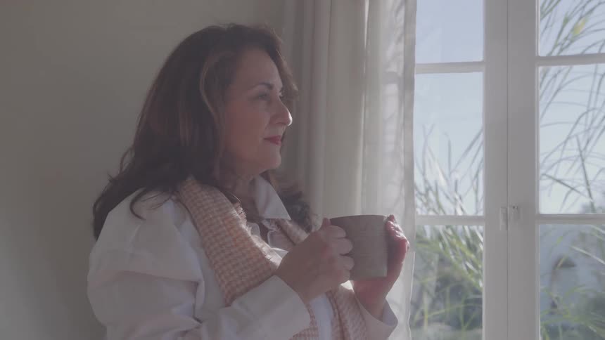 Woman holding a mug while looking out the bright window at the landscape