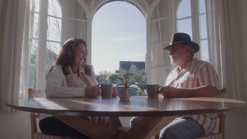 An older woman with long brown hair and an older man wearing a hat and striped shirt sit at a wooden table enjoying coffee and conversation by a large arched window with bright sunlight streaming in