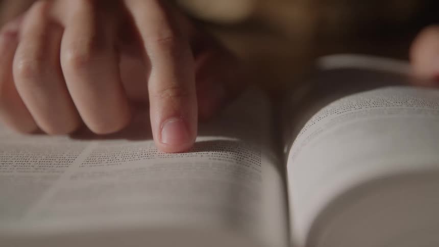 A closeup camera shot shows a hand holding an open book with the index finger moving from left to right across a line of printed text