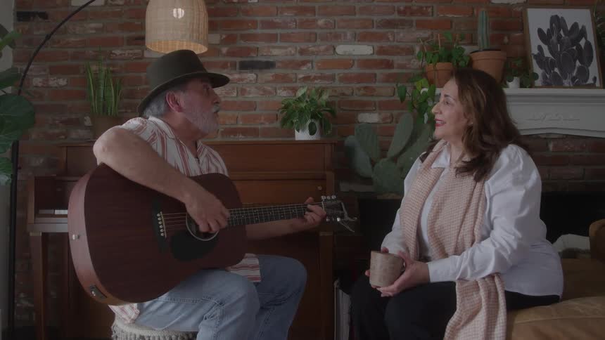 An older man wearing a hat playing an acoustic guitar and singingtalking to an older woman holding a mug as they sit across from each other in a room with a brick wall