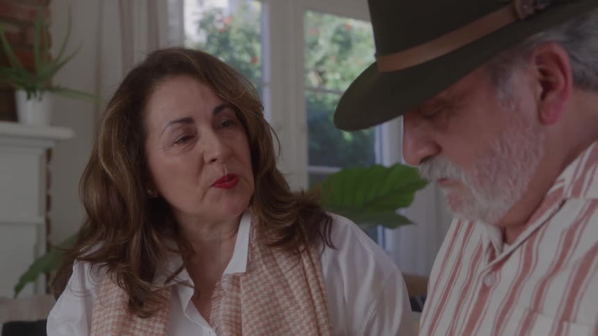 The face of an older woman with long brown hair talking and smiling at an older man wearing a dark hat