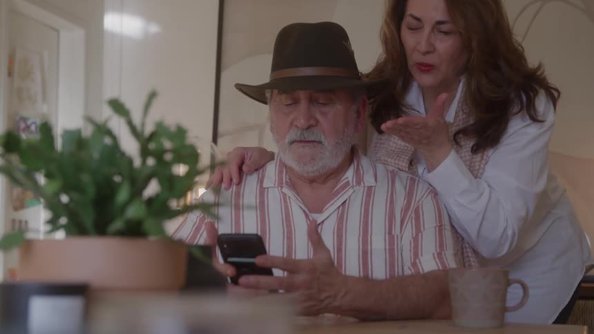 An older man wearing a hat sitting at a table and looking at a black smartphone while an older woman leans over him places a hand on his shoulder and gestures with her other hand