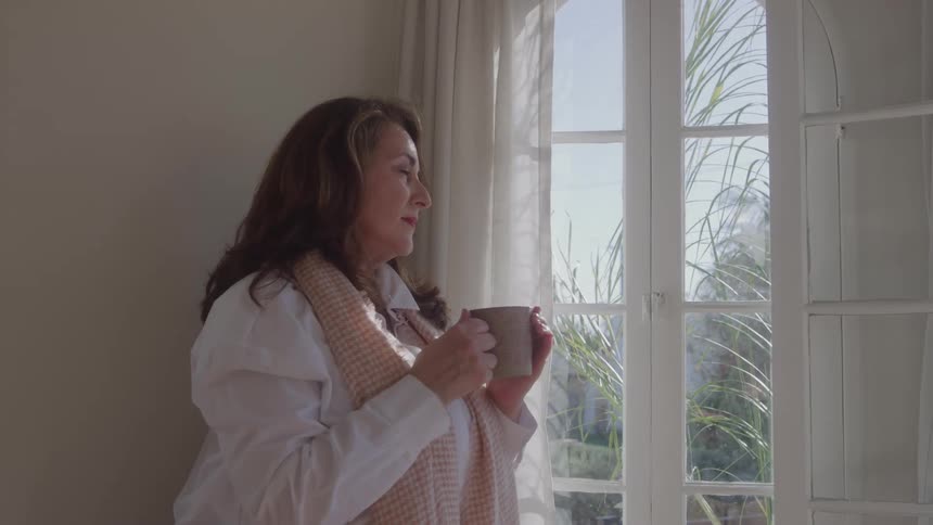 A mature woman with a scarf and holding a tea cup looking outdoors through a window