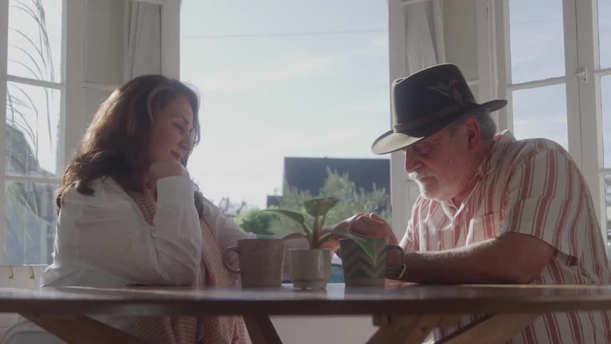 Man kisses the hand of a smiling woman as they sit at a sunlit table