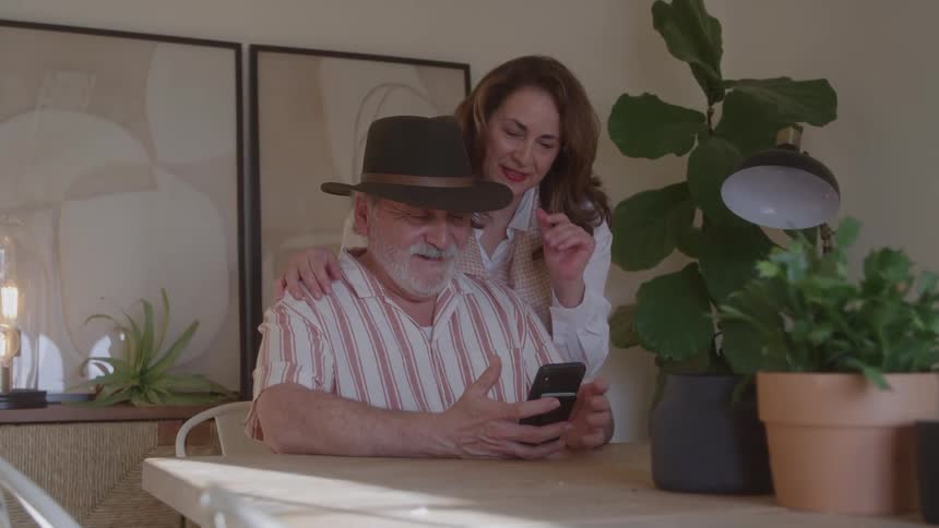 Smiling couple interacting with a smartphone at a table lit by natural light