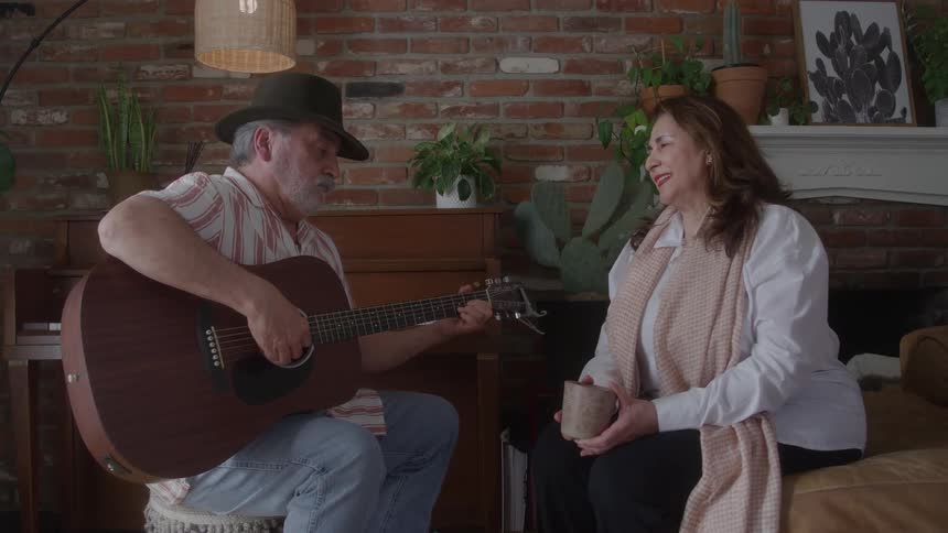 A middleaged couple spends midday together the man wearing a hat and sporting a white beard sits on a chair playing a guitar while the woman sits on the edge of the bed smiling singing and holding a mug