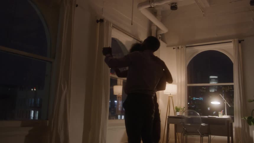 The silhouettes of a man and a woman are seen slowly dancing and embracing in a dimly lit loft apartment with tall arched windows looking out onto the city skyline at night