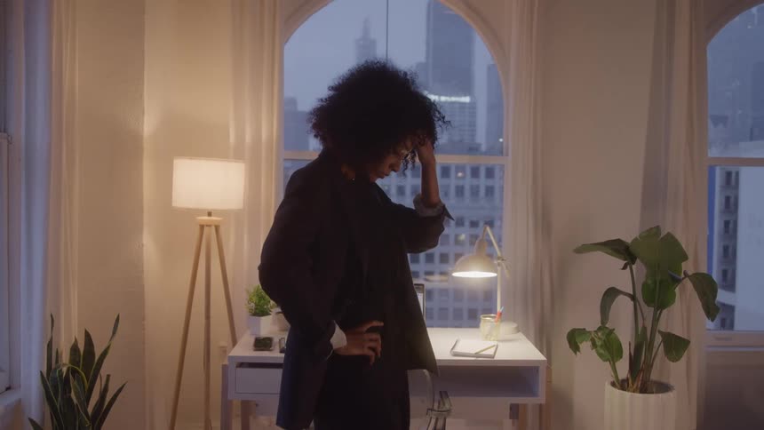Woman standing beside a desk with a thoughtful expression in a softly lit room at dusk