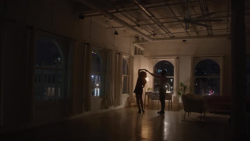 The silhouettes of a man and a woman are seen slowly dancing in a dark wide loft apartment with arched windows overlooking the city lights at night