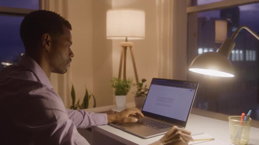 Man works at night writing in a notebook beside a laptop on a lit desk