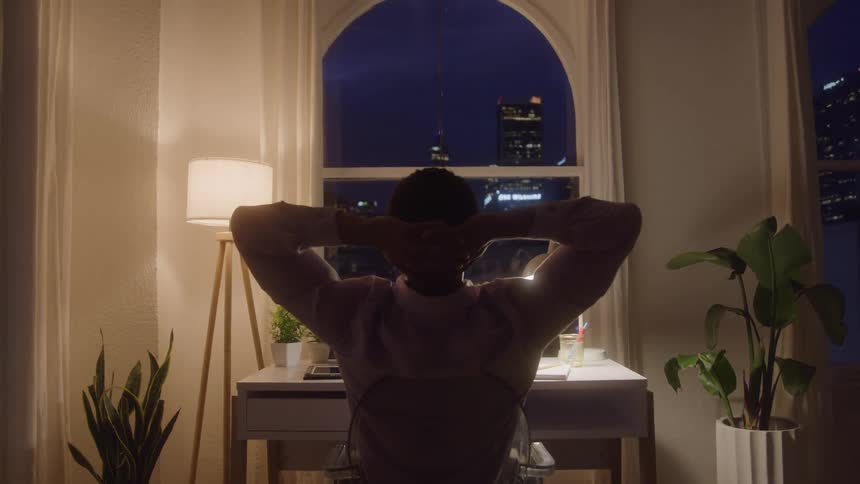 A man sits at a desk in a dimly lit room facing away from the camera and toward an arched window overlooking a glowing city skyline at night