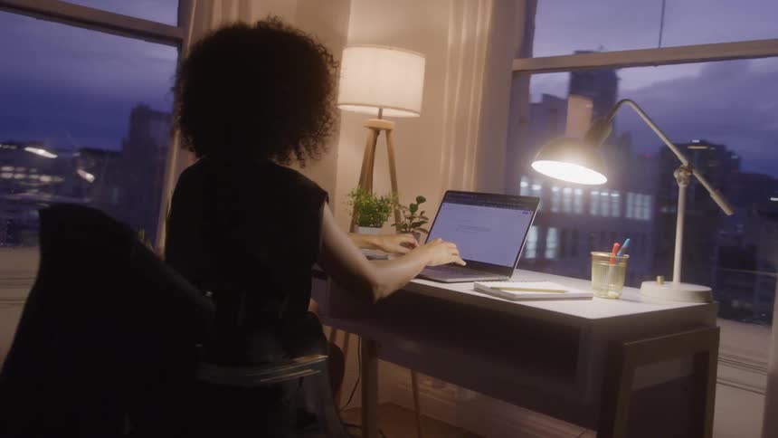 A person with curly dark hair walks from an interior space to an arched window overlooking a city skyline at dusk sits down at a desk with a task lamp and begins typing on a laptop computer
