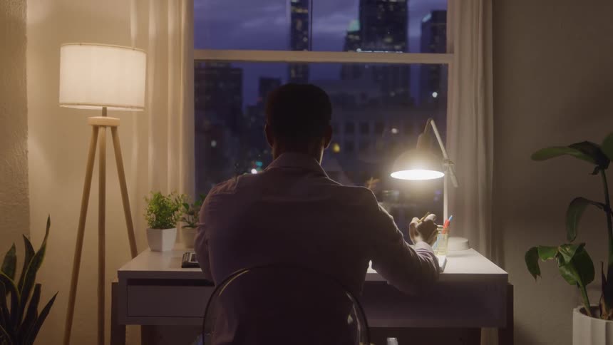 Man seated at a desk writes at night facing a window lit by the city