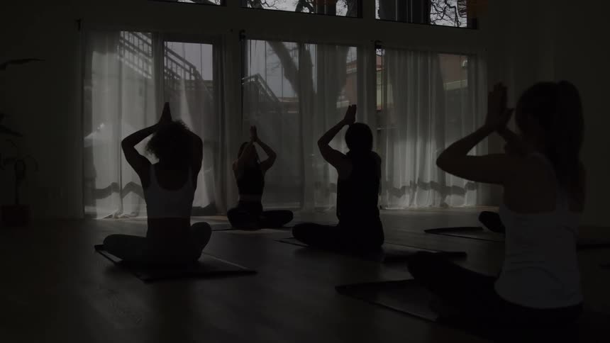 Silhouetted figures perform yoga in a dimly lit studio with large windows and sheer white curtains transitioning from arms raised to hands on their heads