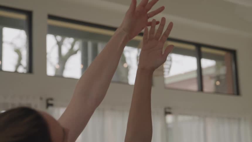The video shows two women in a bright space stretching or practicing yoga one woman with light brown curly hair raises her arm while the other woman gently places her hands on the raised arm seemingly guiding the stretch or pose