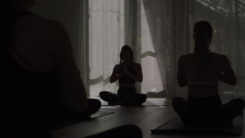 Silhouetted figures of women meditate in a bright yoga studio sitting cross legged on mats with their hands pressed together