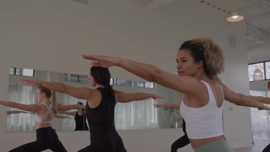 In a bright studio with large mirrors a diverse group of women performs warrior pose during a yoga class