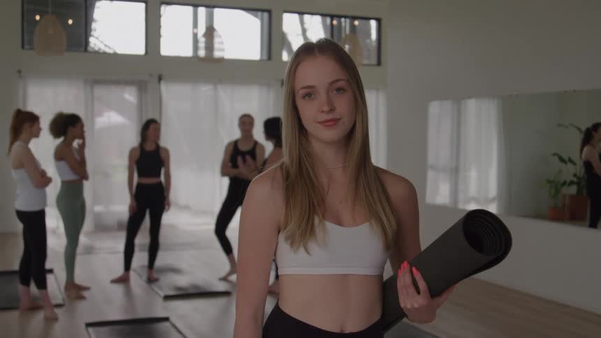 The video shows a young woman in a sports bra and leggings holding a rolledup yoga mat and smiling directly at the camera in the background other women are scattered around a bright studio standing on their mats and preparing for a yoga or fitness cla