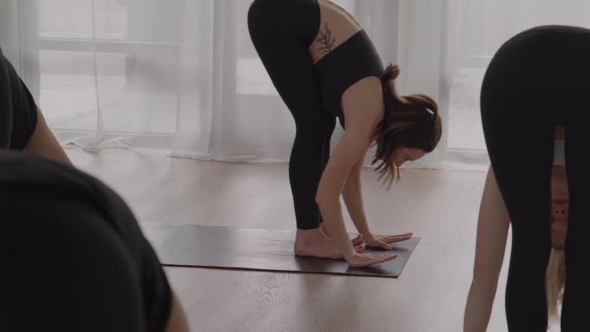 The video features a group of people primarily seen from the back performing a standing forward bend uttanasana on yoga mats in a bright studio they are stretching and moving through a yoga sequence as part of a class