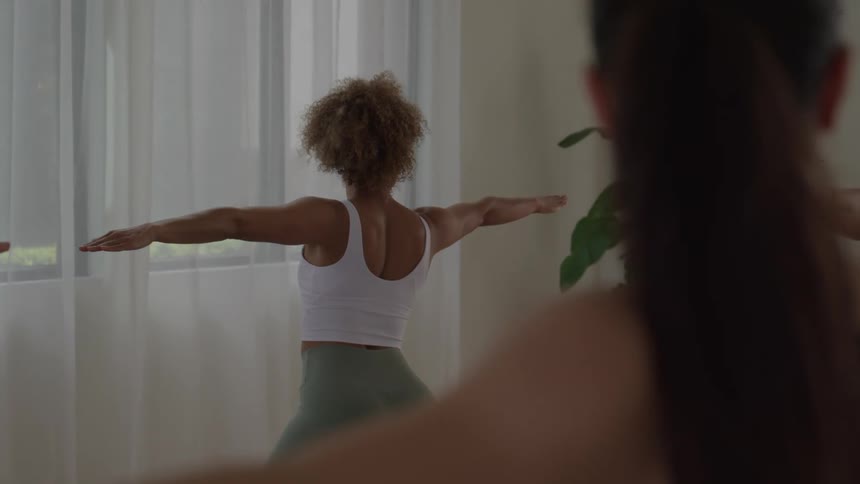From behind a woman with curly hair performs a yoga pose with outstretched arms facing a bright window in a studio with other blurred figures
