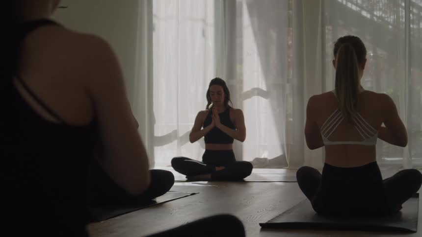A group of women in a bright yoga studio sits on mats in a cross legged meditative pose with their hands pressed together in front of their chests