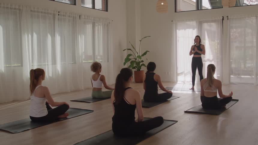 The video shows a yoga class in a bright modern studio with several students seated on mats in a crosslegged position a yoga instructor stands in front of them talking and giving directions to the class