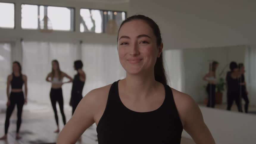 A smiling woman wearing a black tank top stands confidently in the foreground looking directly at the camera in the background of the bright studio several other women in athletic wear are standing on mats suggesting the setting is a yoga or fitness c