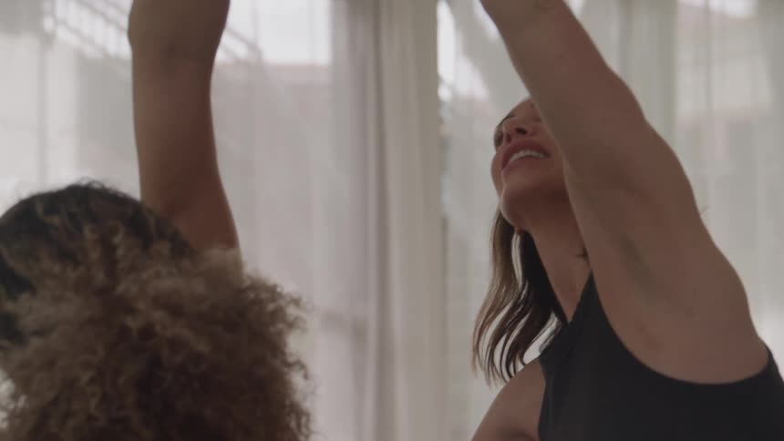 The video shows two women in a bright room doing what looks like a stretching or yoga exercisein the beginning one woman is kneeling with the others hand on her lower back seemingly helping with posture or a stretch then both women raise their