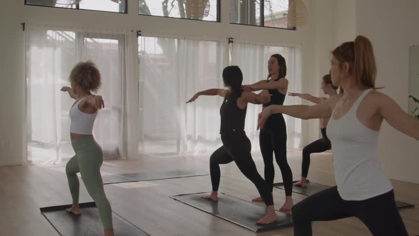 In a bright spacious yoga studio a diverse group of women performs warrior pose extending their arms horizontally