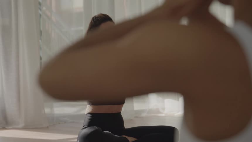 The video focuses on a woman sitting in a crosslegged pose sukhasana in a bright studio likely at the start or end of a yoga sessionshe inhales stretching her arms overhead then brings her hands down to her chest in prayer position an