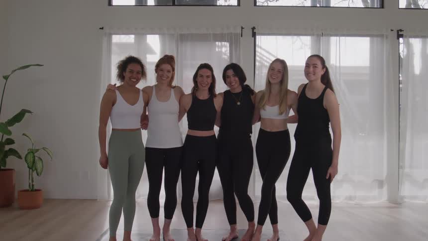 A diverse group of six smiling women in activewear stands arm in arm in a bright yoga studio posing for a group picture