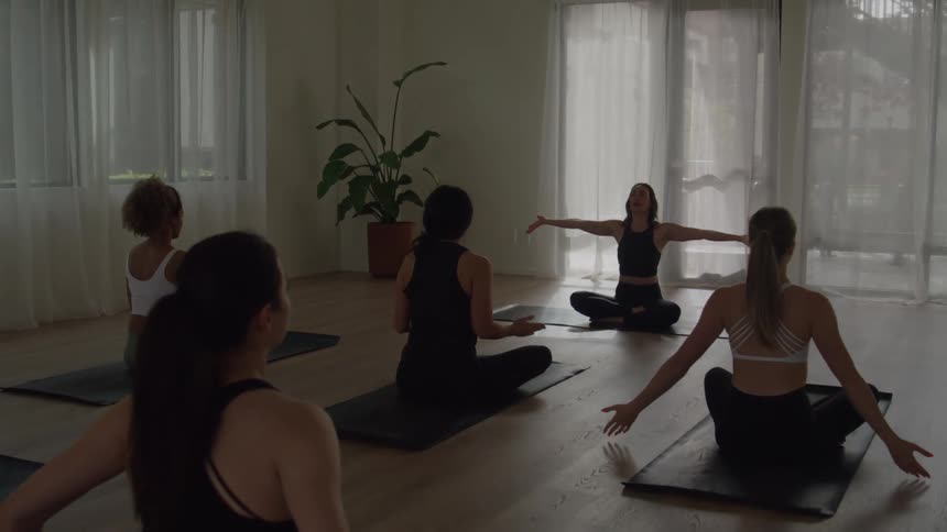 The video shows a yoga class with five women seated in a circle on their mats in a bright modern studioled by the instructor in the center they transition from holding their hands in prayer position anjali mudra to extending their arms ou