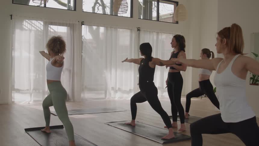 The video features a group of five women in a bright sunlit studio performing a yoga routinethey start by holding the warrior ii pose virabhadrasana ii with arms extended then simultaneously transition by raising one arm overhead whil