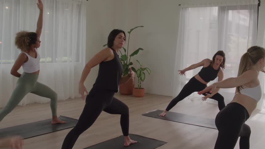 The video shows a group of four women performing yoga poses in a bright spacious studiothey begin in a variation of reversed warrior with one arm extended up then transition into an extended side angle or a similar pose demonstrating fluidit