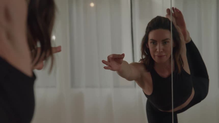 A focused woman in a black yoga outfit holds a challenging yoga pose in a studio with a mirrored wall