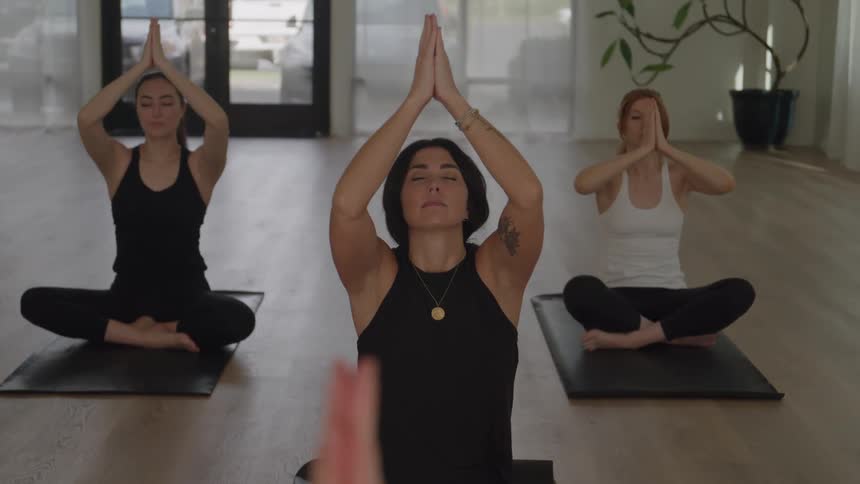 The video shows three women seated in a crosslegged pose sukhasana on yoga mats likely beginning or ending a sessionthey perform a synchronized movement sweeping their arms out raising them overhead to meet in prayer position and then