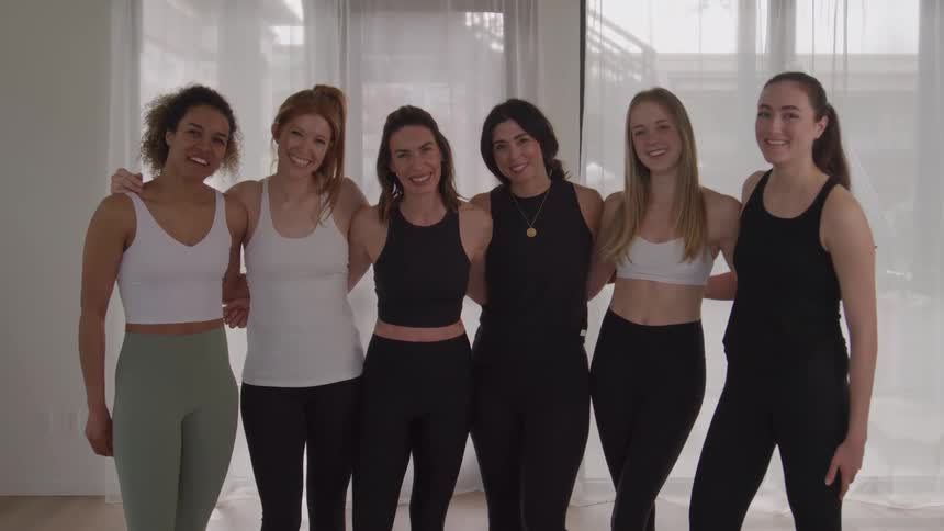 Six women pose side by side in sportswear in a yoga studio