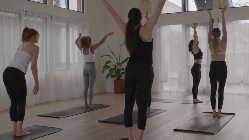 A group of women standing on mats in a room with large windows rising from a deep forward bend to a standing position raising their arms over their heads