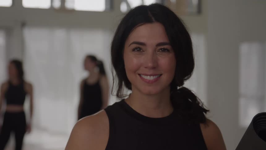 A smiling woman in a black tank top and ponytail holds a yoga mat and looks directly at the camera in a bright studio with other women blurred in the background