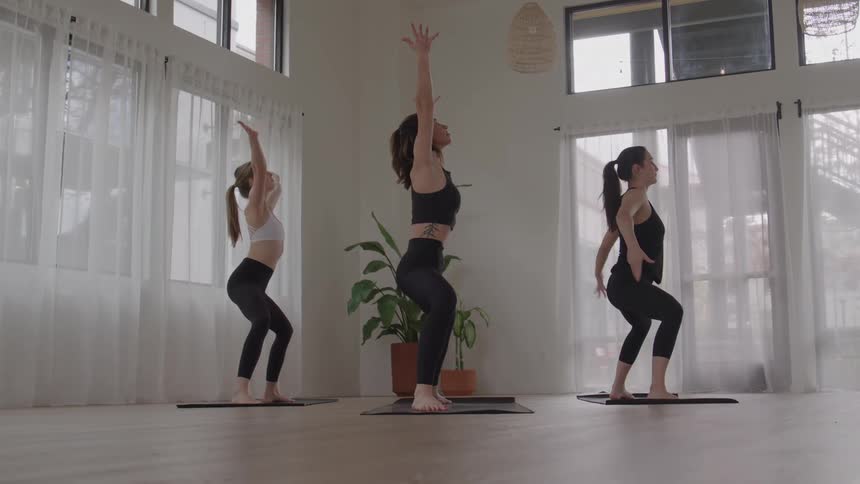 Three people perform chair pose with raised arms in a yoga studio