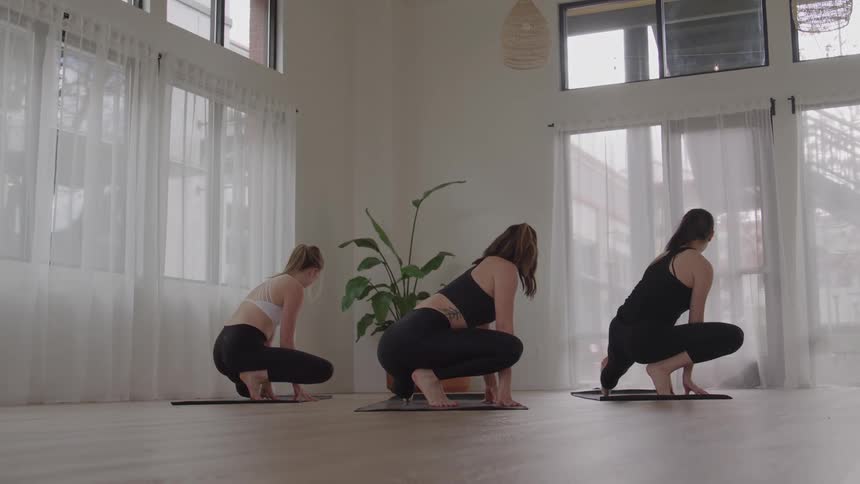 Three young women practice yoga at midday inside an empty room with white seethrough curtains the one in the middle leads the posture changes while the other two follow with a flower pot placed behind them
