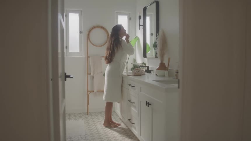 A young woman wearing a white bathrobe does her makeup in front of the bathroom mirror at midday