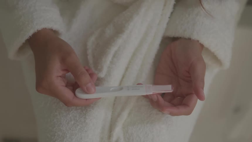 A woman wearing a bathrobe stands in the bathroom holding a urine pregnancy test