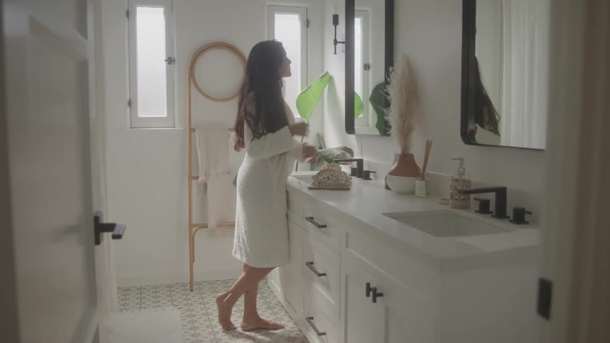A young woman wearing a bathrobe stands in front of the bathroom mirror at midday fixing her hair