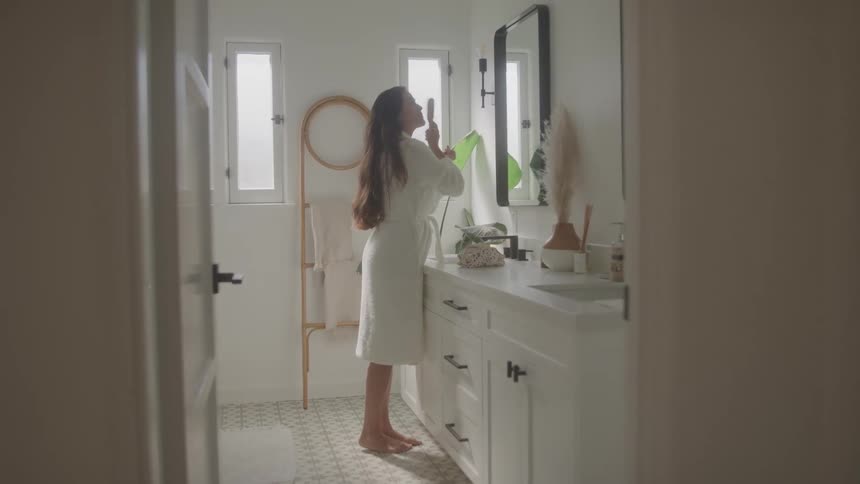 Woman in a white robe stands combing her hair in front of the mirror in the bathroom