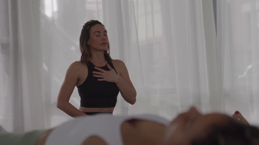 Women in gym closes lying on the floor of a gym while another is sitting with her hand on her chest and her eyes closed doing breath work then tilts her head forward and puts her hands together in a prayer stance