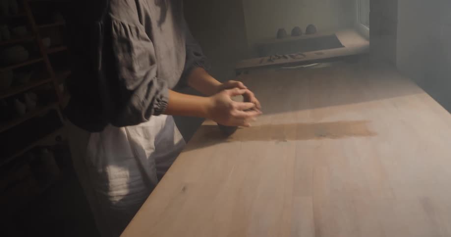 The video shows a woman standing at a wooden counter in a dimly lit room intensely focused on her taskshe is vigorously kneading or working a dark lump of material with her hands possibly clay for pottery or dough for baking next to a sunlit