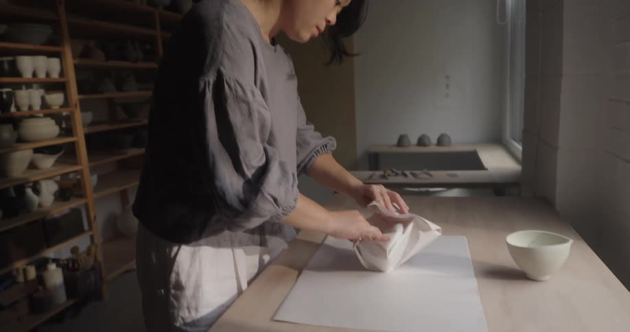 A young asian woman wrapping a ceramic bowl with paper sheets on a tabletop in a pottery room during daytime