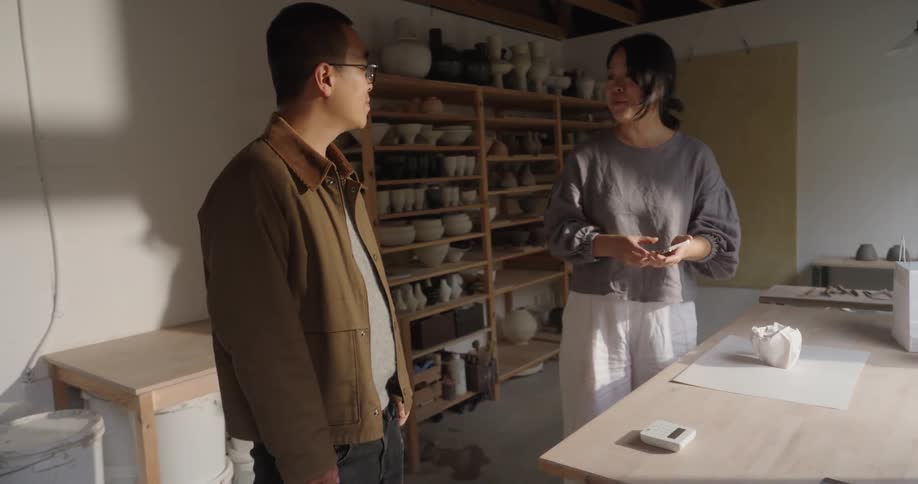 A man in a brown jacket entering a room with exposed beams and shelves full of ceramic vessels where a woman greets him handles a piece of pottery and then the two people stand facing each other while operating small black electronic devices in their h