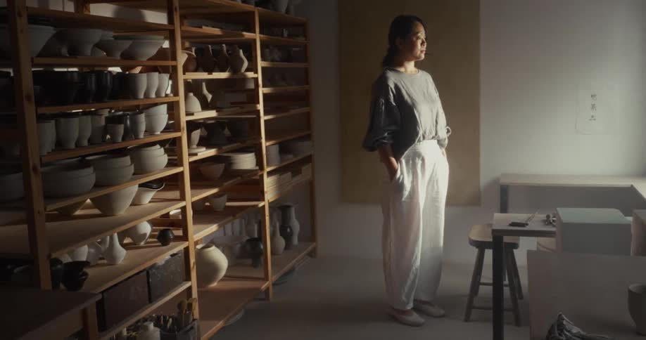 A woman stands contemplatively in a sunlit pottery studio surrounded by shelves filled with various ceramic pieces